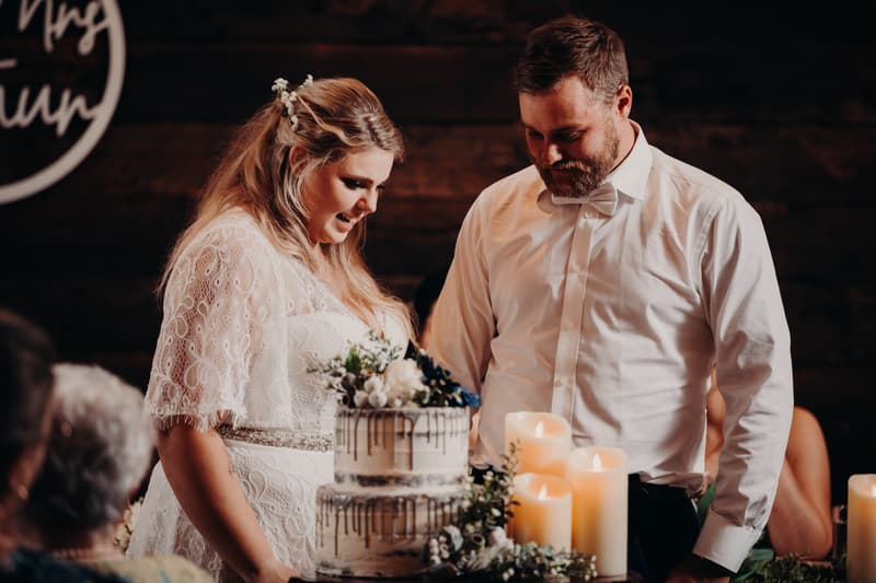 Bride Annie and groom Trent stand together behind a two-tiered wedding cake decorated with flowers and surrounded by lit candles at Ocean View Estates reception stage.
