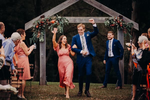 A bridesmaid and a groomsman walk down the aisle arm in arm under a wooden arch decorated with flowers at Yabbaloumba Retreat — By The River, while guests on either side applaud and take photos.