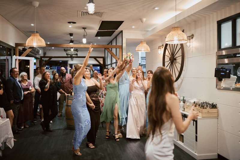 The bride Olivia, with her back to the camera, tosses her bouquet to a group of women guests at the reception stage of White Horse Ranch. Several women in dresses reach up to catch the bouquet while other guests watch in the background.