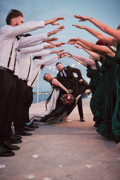 Christopher dips Jayme holding a bouquet as bridesmaids in dark green dresses and groomsmen in white shirts with suspenders form an arch with their arms. Wedding family photos by She Said Yes.