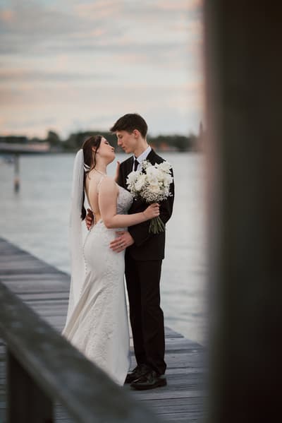 The bride Terion and groom Tyler embrace on a wooden pier, with the bride holding a bouquet of white flowers and the water visible in the background. Best Brisbane wedding photographer by She Said Yes.