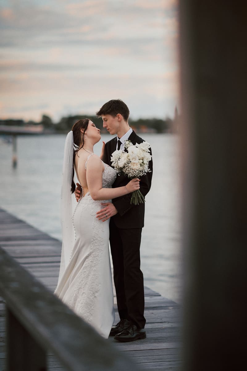 The bride Terion and groom Tyler embrace on a wooden pier, with the bride holding a bouquet of white flowers and the water visible in the background. Full day wedding photography by She Said Yes.