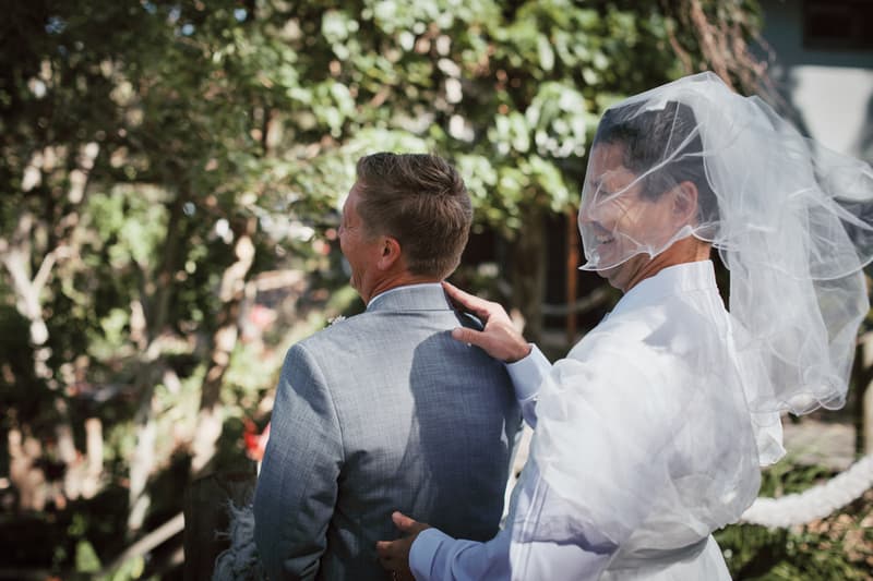 The bride, Jheng, wearing a veil and white dress, touches the shoulder of the groom, Dane, who is facing away and wearing a light grey suit, during a moment outdoors at Sandstone Point Hotel.