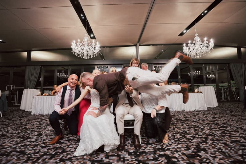 At the Sandstone Point Hotel — The Pumicestone Room, the bride Morgan in a white wedding dress and the groom Joel in a light suit sit among guests while a man in a dark shirt and white pants is held horizontally above them in a playful group pose under chandeliers.