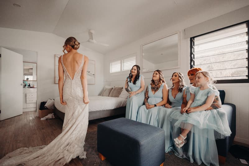 The bride stands with her back to the camera showing her lace wedding gown while five bridesmaids and a flower girl in matching light blue dresses sit on a couch in a bedroom.