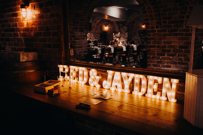 Illuminated marquee letters spelling 'RED & JAYDEN' on a wooden table at Sandstone Point Hotel — The Cellar, with a mirror reflecting a brick-walled dining area set for a reception.