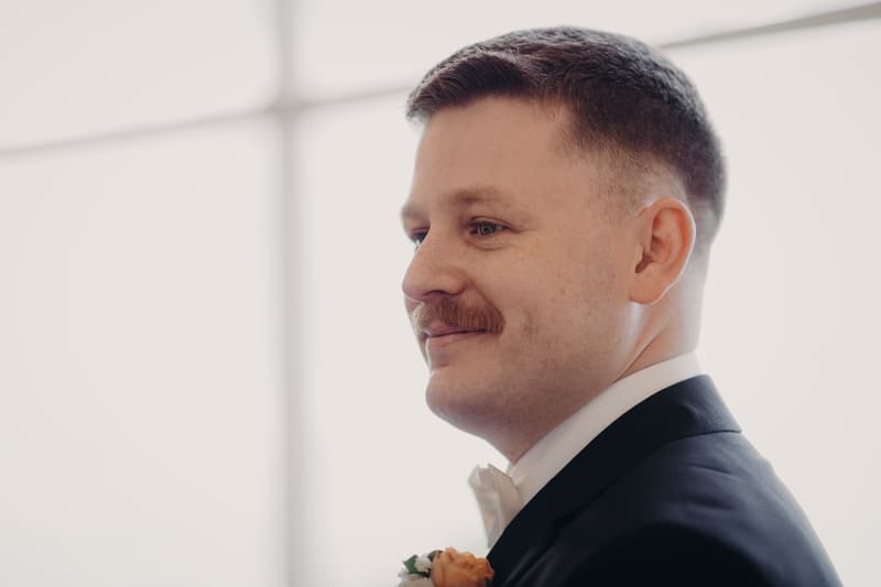 The groom Liam in a black suit and white bow tie with a boutonniere, standing indoors at Tiffany's Maleny — Chapel.
