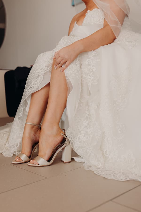 The bride sits indoors wearing a white lace wedding dress and beige high-heeled sandals, with her hands resting on her knee.