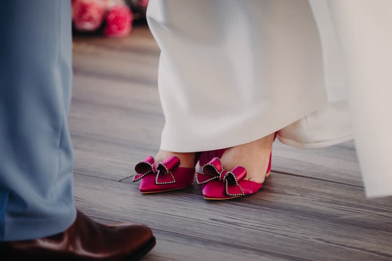 The bride wears bright pink heels with large bows, standing on a wooden floor at Sandstone Point Hotel — Pavilion, with the groom's brown shoe and light blue pants partially visible.