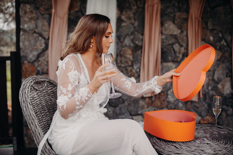 The bride Chelsea in a white lace wedding dress sits on a wicker chair at Preston Peak Winery Functions Centre, holding a glass of champagne and lifting the lid off a heart-shaped orange polka dot box.