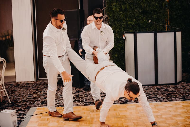 Two groomsmen hold the legs of a man in a white shirt and light pants as he walks on his hands on the dance floor at Sandstone Point Hotel — Pumicestone Room during the wedding reception.