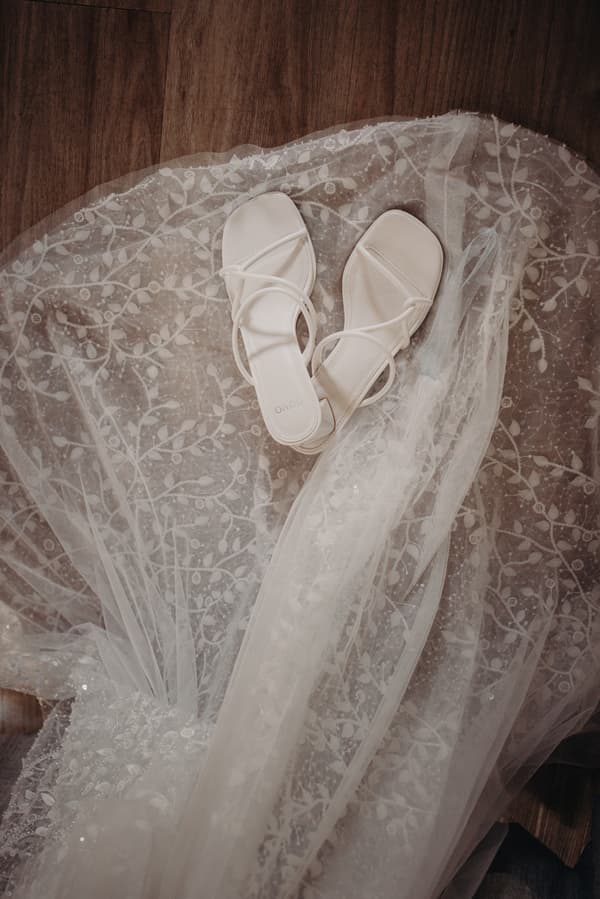 White bridal sandals placed on a lace-patterned bridal veil or wedding dress fabric on a wooden floor.
