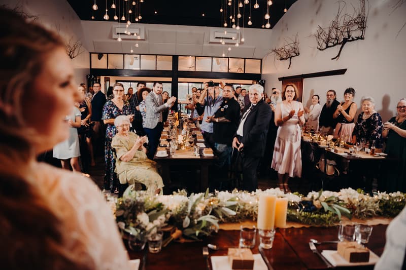 Guests at Ocean View Estates stand and applaud during the wedding reception, with the bride Annie partially visible in the foreground.