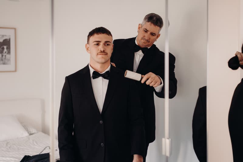 Kyle, the groom, stands while an older man in a tuxedo uses a lint roller on his jacket in a bedroom setting.