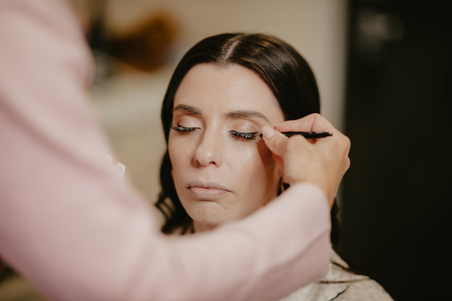 A bride having eyeliner applied to her closed eyes by a makeup artist during preparation.