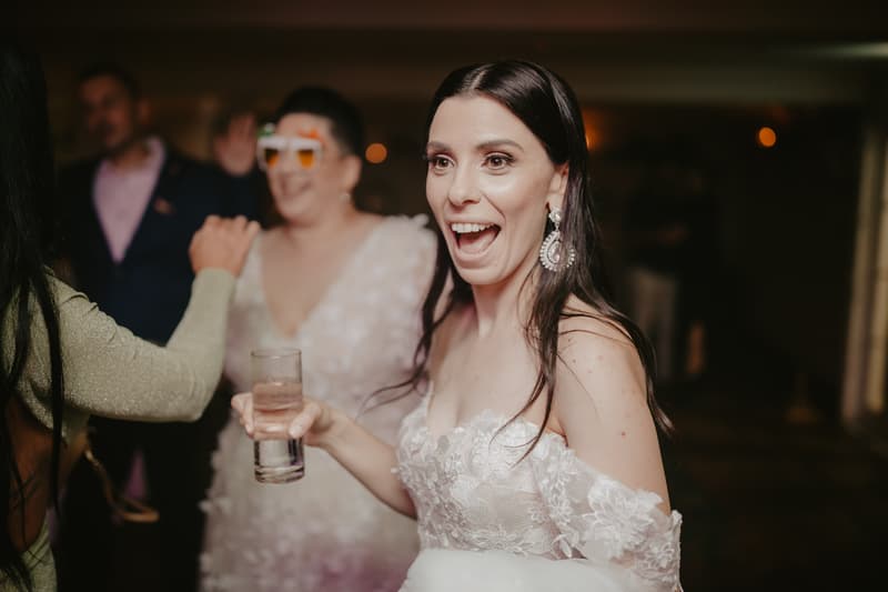 Bride in a white lace wedding dress holding a glass, smiling with guests in the background.