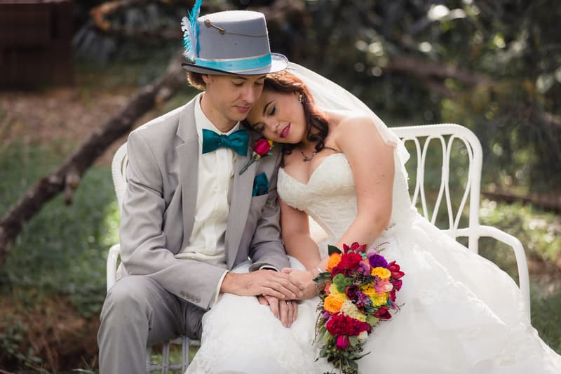 Bride Holly in a white wedding gown holding a colorful bouquet leans her head on groom Paul's shoulder as they sit together on a white bench outdoors at Kwila Lodge.