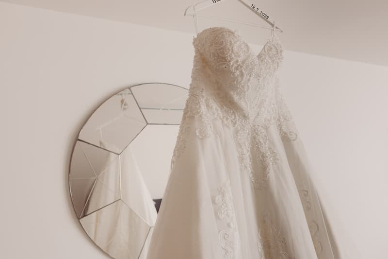 Emily's wedding dress with intricate lace detailing hangs on a personalized hanger in front of a decorative round mirror.