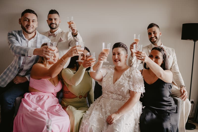 A group of seven people, including a bride in a white floral lace dress, two men in light-colored suits, and four others in formal attire, sit closely together on a couch raising glasses of rosé wine in a toast.