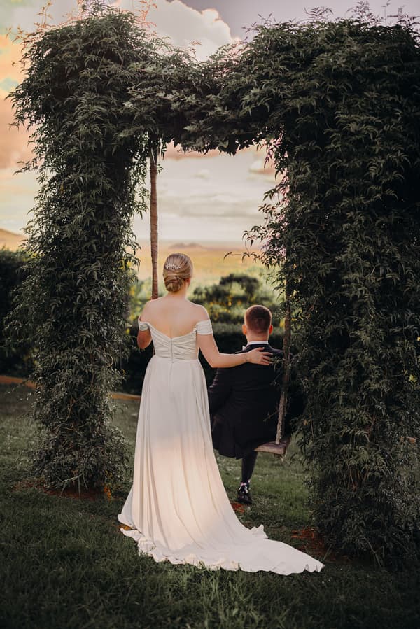 The bride Courtney stands beside the groom Liam who is seated on a swing framed by a leafy arch at Tiffany's Maleny, both facing away from the camera toward a scenic landscape.