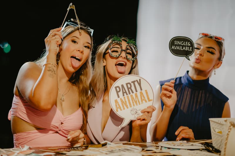 Three women pose at a table with photo booth props, including signs reading 'PARTY ANIMAL' and 'SINGLE & AVAILABLE'. Two women stick out their tongues, and one woman puckers her lips. The background is dark with a white panel on the right.