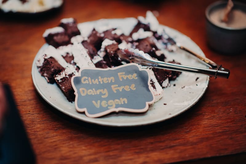 A plate of gluten free, dairy free, vegan brownies with a small sign and serving tongs on a wooden table. Getting ready photos by She Said Yes.