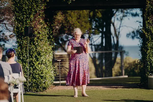 A woman in a patterned dress stands under a rustic arbour at Sandstone Point Hotel, reading from a tablet during the wedding ceremony while guests are seated nearby.
