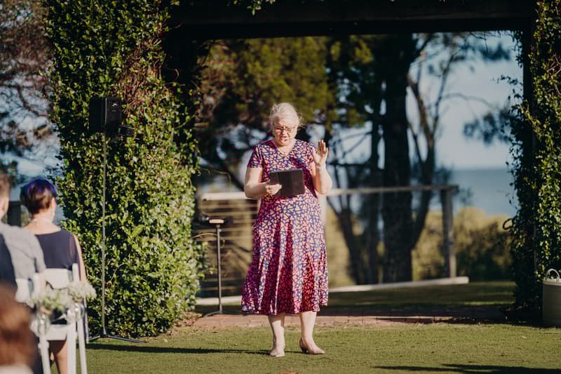 A woman in a patterned dress stands under a rustic arbour at Sandstone Point Hotel, reading from a tablet during the wedding ceremony while guests are seated nearby.