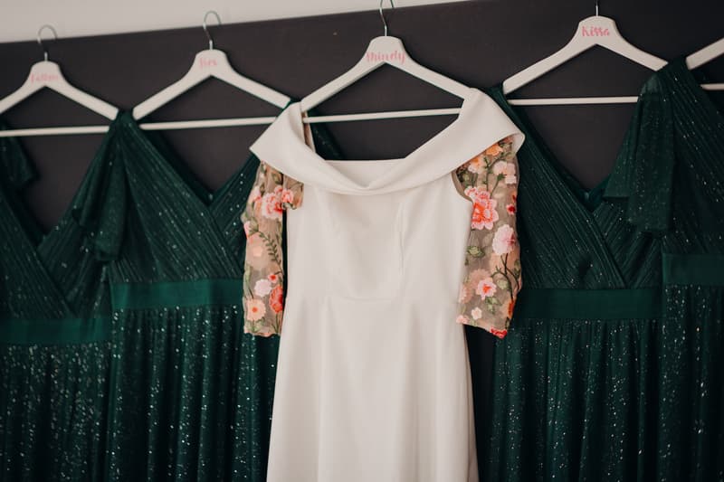 The bride's white wedding dress with floral embroidered sleeves hangs on a hanger labeled 'Mindy' alongside three dark green bridesmaids' dresses on hangers labeled 'Kissa', 'Ben', and 'Lillian'.