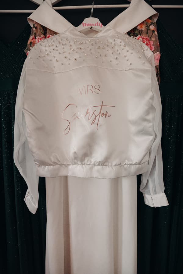 The bride's white satin jacket with pearl embellishments and the text 'MRS Searston' hangs on a personalized hanger labeled 'Mindy' over a white wedding dress.