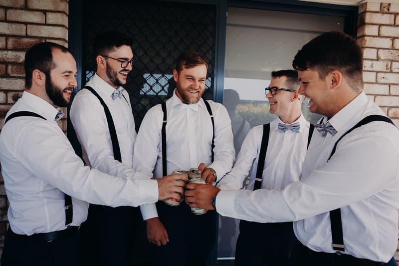 The groom and four groomsmen in white shirts, bow ties, and suspenders toast with canned drinks outside a brick building at Ocean View Estates.