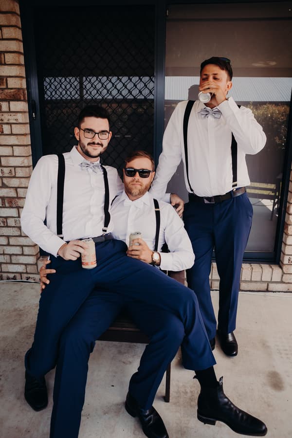 Three groomsmen wearing white shirts, navy pants, gray bow ties, and black suspenders pose casually outside Ocean View Estates, two sitting on a chair holding cans and one standing drinking from a can.
