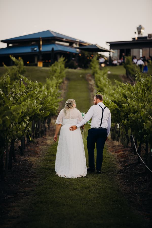 The bride Annie and groom Trent walk hand in hand down a grassy path between rows of grapevines at Ocean View Estates during their couple portraits session.