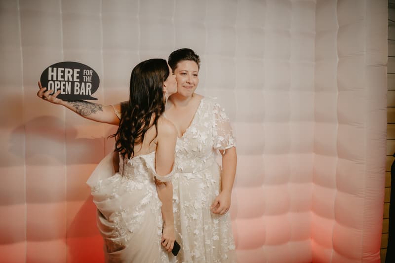 Two brides in white wedding dresses pose in front of a white inflatable backdrop; one bride kisses the other on the cheek while holding a sign that reads 'HERE FOR THE OPEN BAR'.