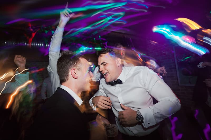 Two men in formal attire dance and sing energetically at the reception in Sandstone Point Hotel — The Cellar, surrounded by colorful party lights and other guests.