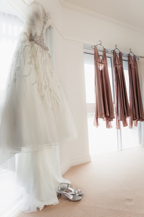 The bride's wedding dress is hanging near a window at Novotel Sunshine Coast Resort with silver bridal shoes placed on the floor beneath it, and three bridesmaids' dresses are hanging on a curtain rod by the window.