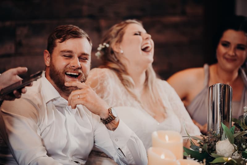 The groom Trent and the bride Annie laugh together at the reception stage at Ocean View Estates, accompanied by a bridesmaid.