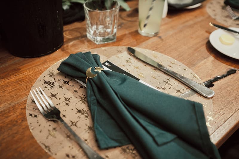 Close-up of a wooden table setting at Sandstone Point Hotel — The Cellar with a gold star-patterned placemat, a dark green napkin held by a moon and star napkin ring, a fork and knife, a glass, and a plate with butter.