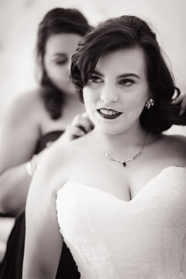 The bride Holly is having a necklace fastened by a bridesmaid during bridal preparations.