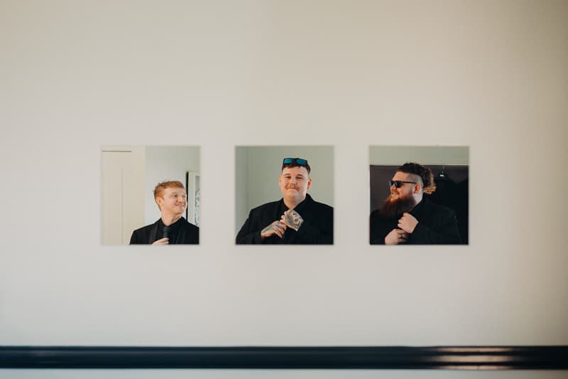 Three groomsmen are individually reflected in three square mirrors mounted on a white wall at Ocean View Estates.