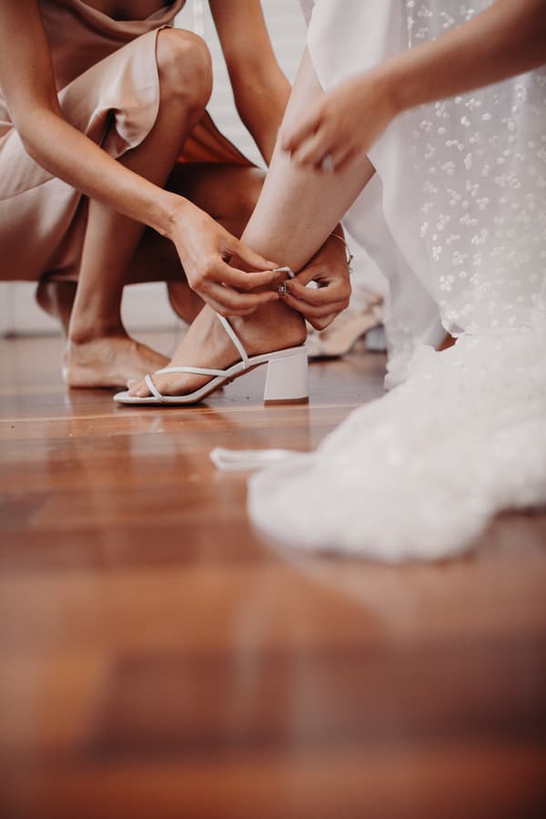 A bridesmaid helps the bride fasten a white sandal on her foot while the bride wears a white wedding dress at Preston Peak Winery Functions Centre.
