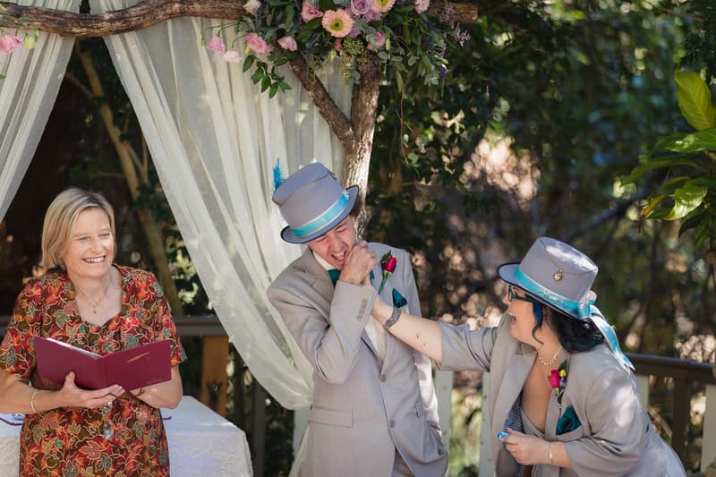 The groom playfully kisses the bride's hand during the ceremony at Kwila Lodge, while the celebrant smiles holding a book.