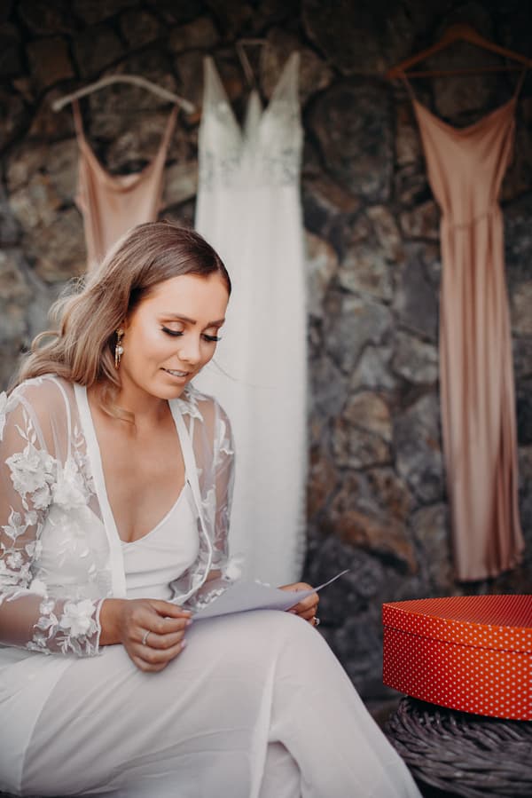 The bride Chelsea sits reading a note with her wedding dress and two bridesmaids' dresses hanging on a stone wall behind her at Preston Peak Winery Functions Centre.