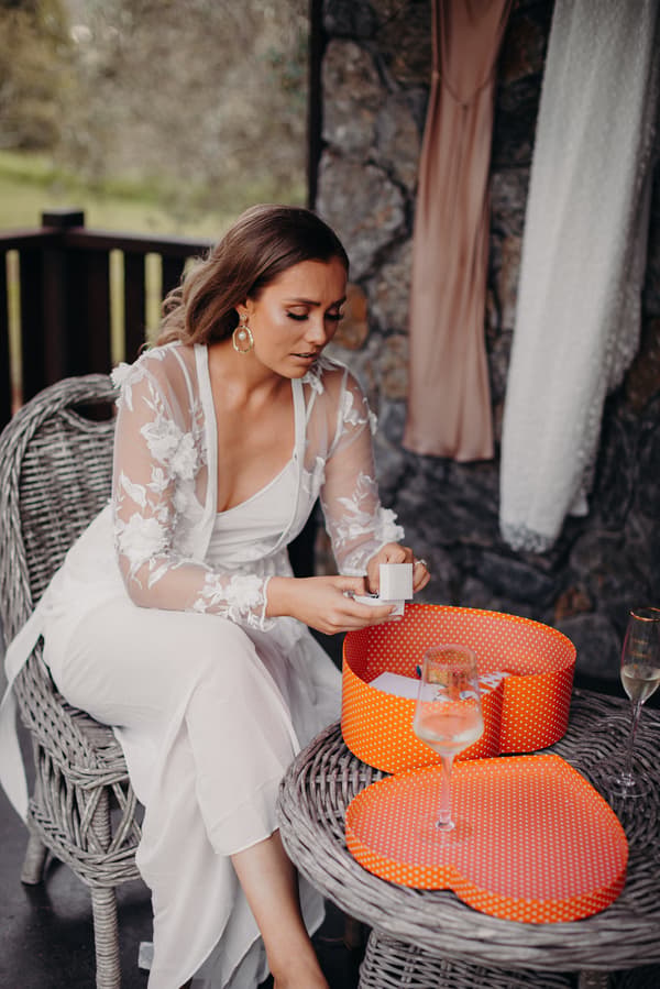 The bride Chelsea sits on a wicker chair at Preston Peak Winery Functions Centre, holding an open ring box with a heart-shaped orange gift box on the table beside her.