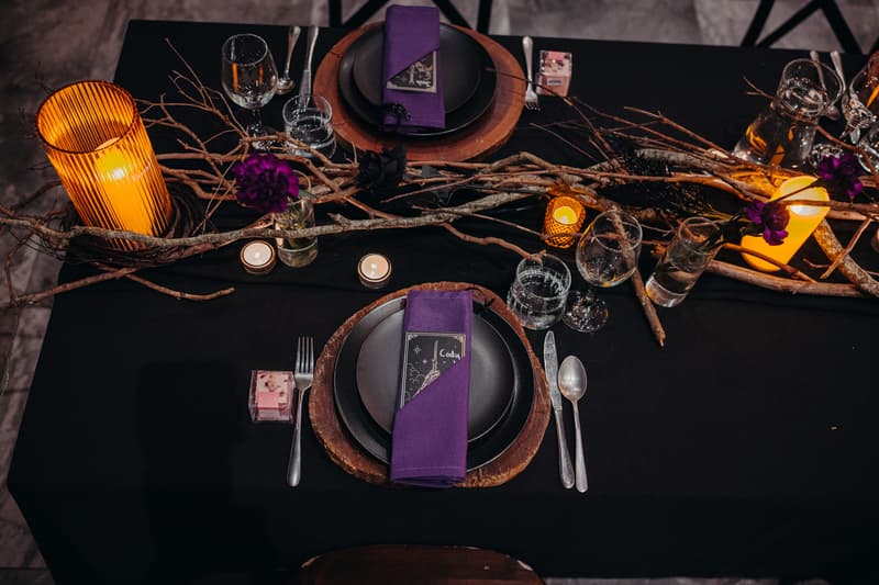 Table setting at Ocean View Estates — Function Room with black tablecloth, wooden chargers, black plates, purple napkins, glassware, candles, and rustic branch centerpiece with purple flowers.