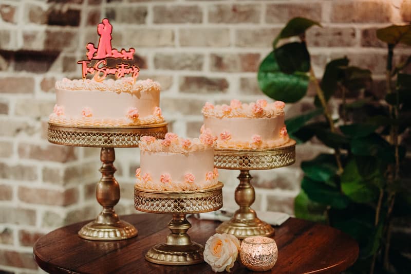 Three pink frosted wedding cakes with small flower decorations are displayed on ornate gold cake stands on a wooden table at Sandstone Point Hotel — Cellar. A pink rose and a lit candle in a glass holder are placed on the table beside the cakes against a brick wall background.