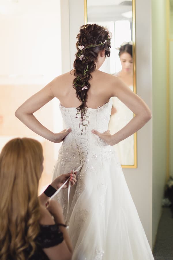 The bride stands with hands on hips facing a mirror while another woman laces up the back of her wedding dress indoors.