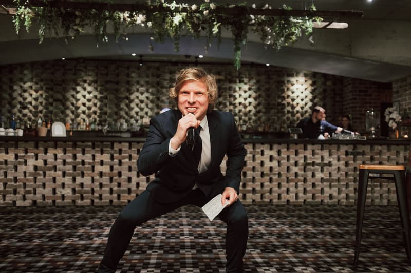A man in a dark suit holding a microphone and a piece of paper is crouching and speaking at the reception stage inside Sandstone Point Hotel — The Cellar, with a bar and two bartenders in the background.