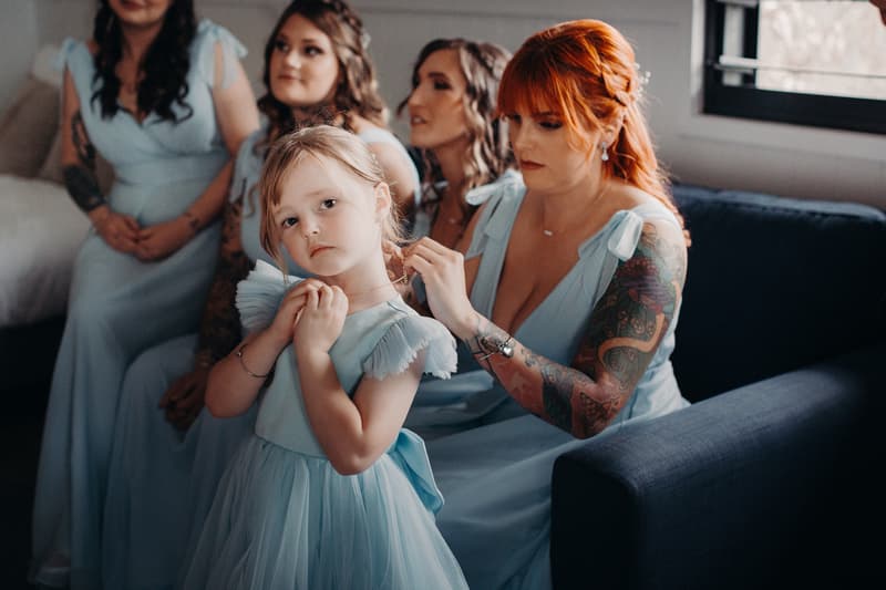 Bridesmaids and a flower girl in light blue dresses sit on a couch inside a room, with one bridesmaid helping the flower girl with a necklace.