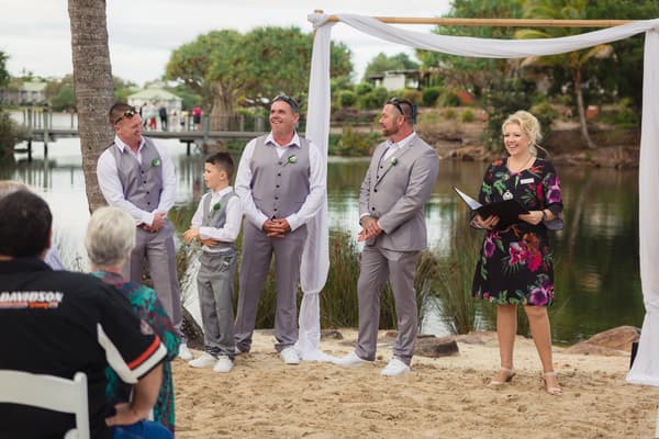 The groom and groomsmen stand under a decorated arch by the water at Novotel Sunshine Coast Resort during the ceremony, with the celebrant holding a book and guests seated in the foreground.